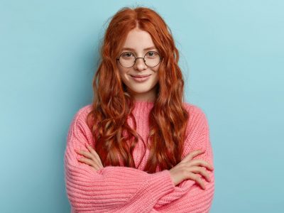 Satisfied ginger charming woman with curly hair, wears transparent glasses, keeps arms folded over chest, has pleasant talk with contemporary, wears oversized clothes, isolated over blue background